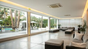 A modern interior with a pool view, large windows, and a seating area with a sofa and ottoman. - Villa Carlino White (Mascali)