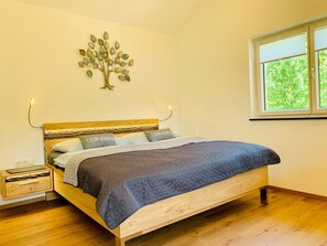 Een slaapkamer met een bed, nachtkastjes, een decoratie van een boom aan de muur en een raam met uitzicht op groen.