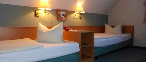 Habitación de hotel con dos camas individuales, cada una con cabecero de madera, ropa de cama blanca y una mesita de noche pequeña. Hay dos lámparas fijadas a la pared que proporcionan luz, y una pequeña estantería entre las camas.