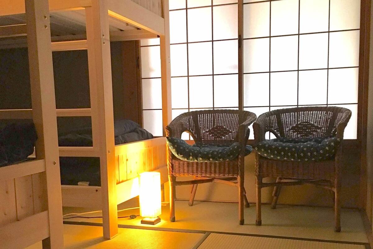 Una habitación con una cama litera, dos sillas de mimbre, una lámpara de pie y un separador japonés.