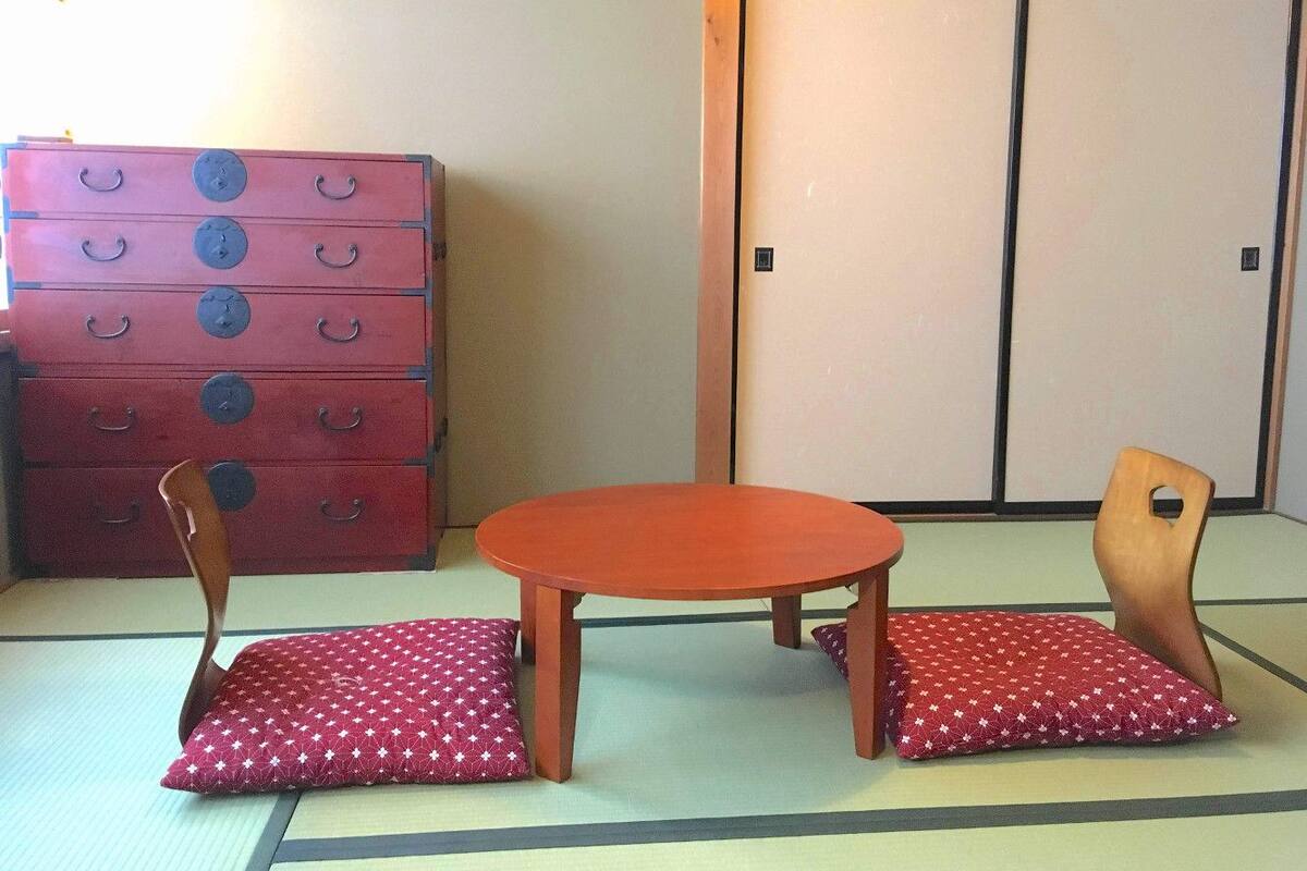 Una habitación japonesa tradicional con suelo de tatami, una mesa de madera baja, dos sillas de madera con cojines y un cofre de almacenamiento rojo.