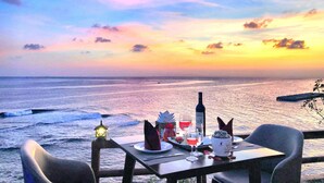 Mesa para dos con botella de vino, copas y un postre, con vista a una playa al atardecer.