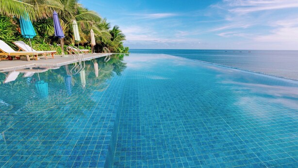 Una piscina infinita de color azul intenso, con sillas de descanso y sombrillas, rodeada de abundante vegetación y vistas al océano.