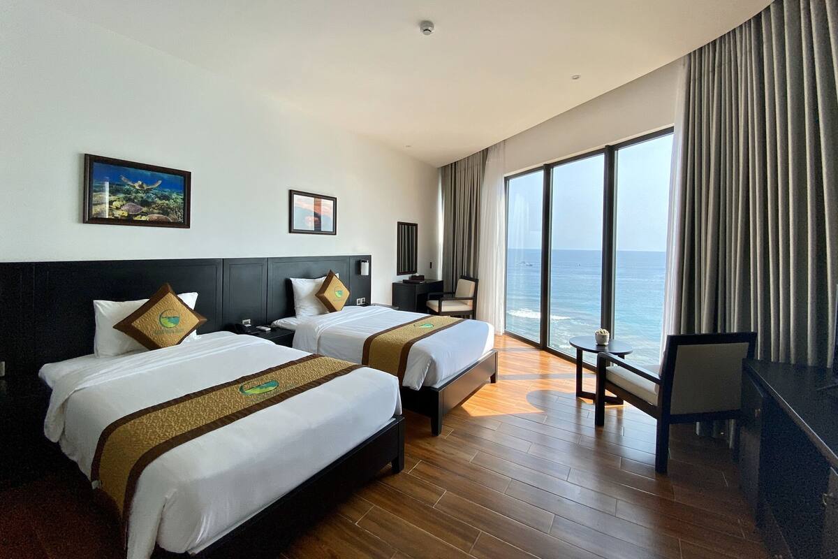 A hotel room with two beds, a desk, a chair, and a large window overlooking the sea.