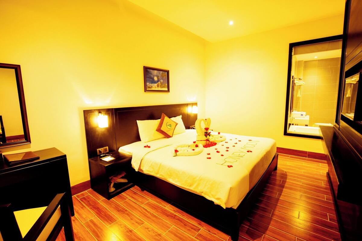 A hotel room with a large bed, bedside tables, a nightstand, a mirror, and a wooden floor.