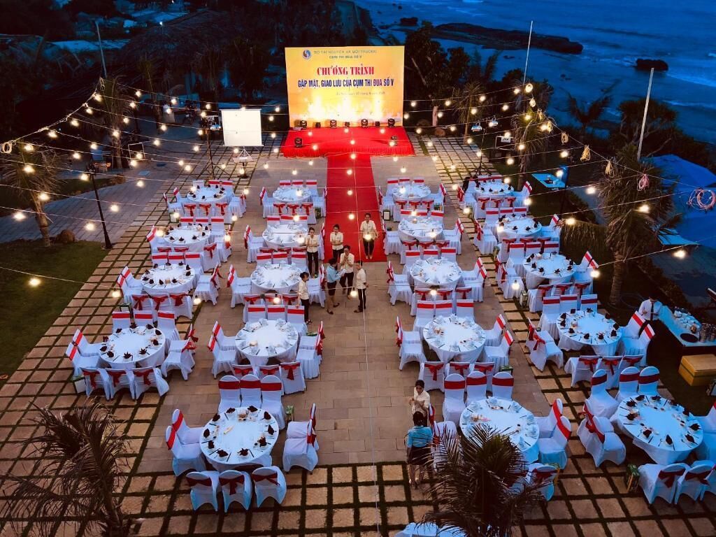 An outdoor event setup with round tables and chairs, a stage with a screen, and string lights.