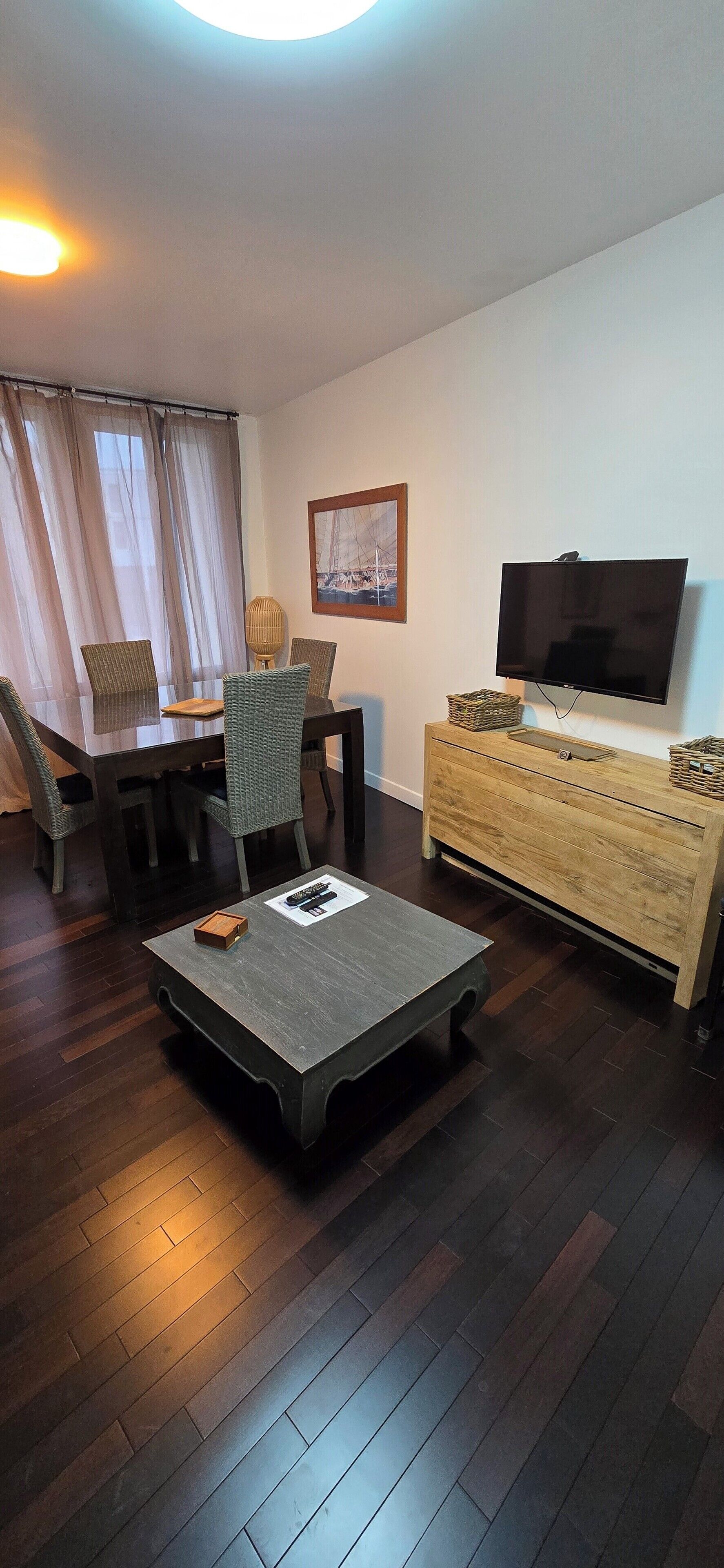 A living room with a dark wood floor, a dining table with chairs, a TV on a wooden cabinet, and a window with curtains.
