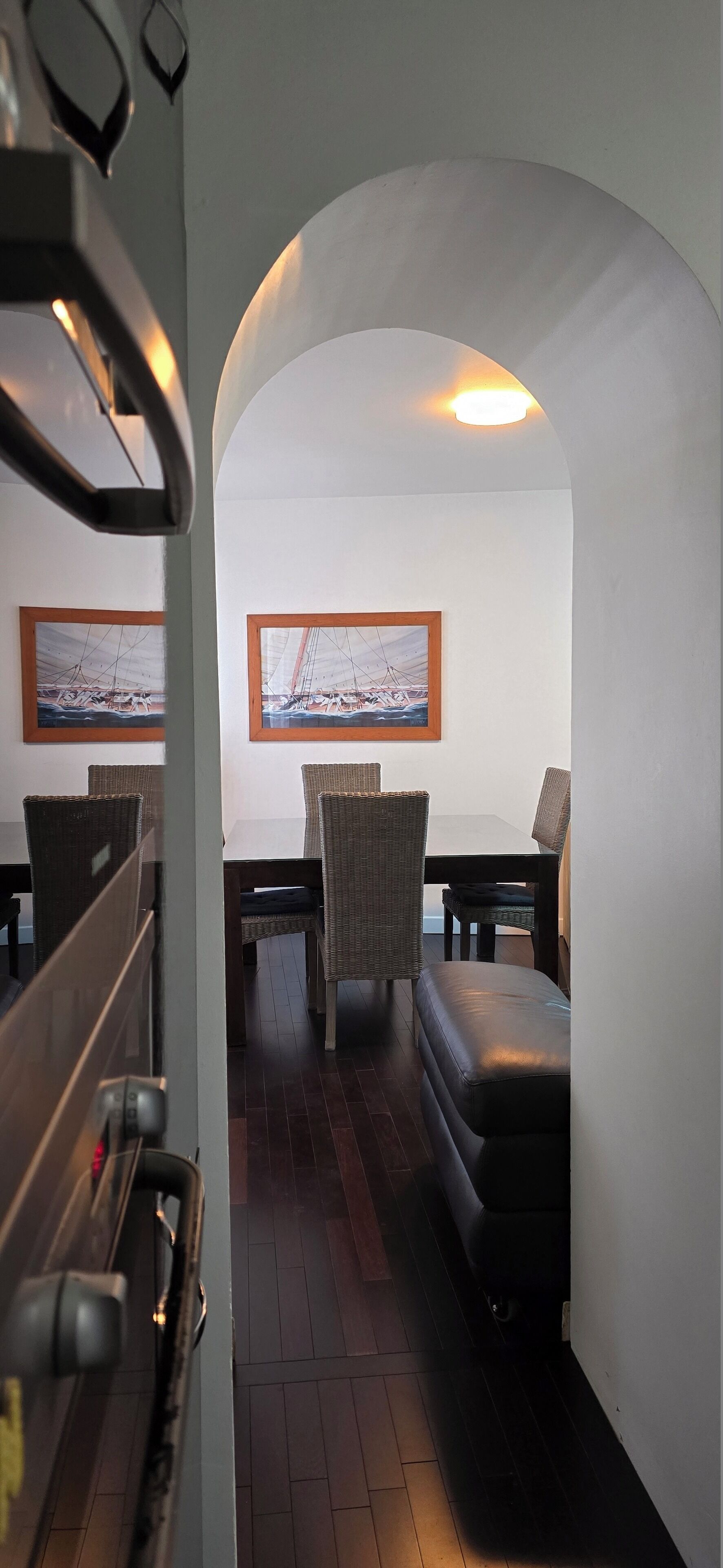 Eine gemütliche Sitzecke mit gepolsterten Bänken, Korbstühlen und einem Tisch. An der Wand hängen gerahmte Bilder, und eine Deckenleuchte ist zu sehen.