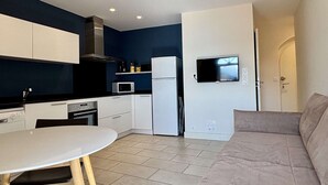 Cozinha compacta com armários brancos, micro-ondas e geladeira. Há uma mesa de jantar com cadeiras e um sofá na sala de estar.