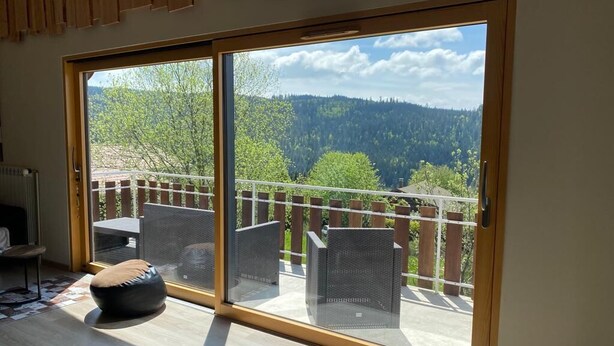 A room with a large sliding glass door leading to a balcony with outdoor seating and a view of a forested area.