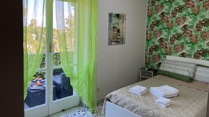 Un dormitorio con una cama, una ventana con cortinas translúcidas, un papel tapiz tropical y un equipo de aire acondicionado.