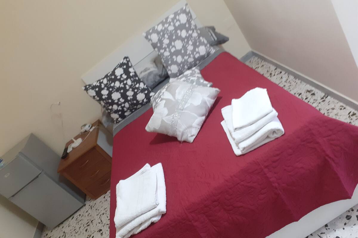 Cama bem arrumada com colcha vermelha, toalhas brancas e almofadas estampadas.