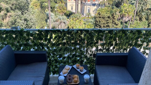 Un balcón con mobiliario de mimbre, una mesa con pasteles y bebidas, y vistas a edificios y árboles.