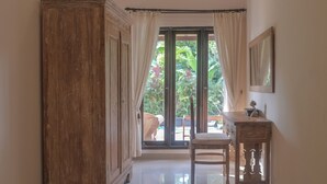 A room with a wooden wardrobe, a desk with a chair, and a large sliding glass door leading to an outdoor area with greenery. - Vila Bukit Sari (Buleleng)