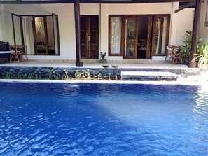 Une piscine bordée de pierres, des chaises longues en bois et une maison avec de grandes fenêtres et une porte en bois.