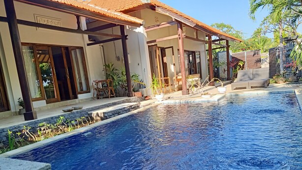 A swimming pool with a tiled deck, surrounded by a covered outdoor area with seating and potted plants. - Vila Bukit Sari (Buleleng)