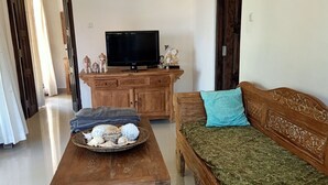 A living room with a wooden coffee table, a sofa with a green throw, a television on a wooden cabinet, and a chandelier. - Vila Bukit Sari (Buleleng)