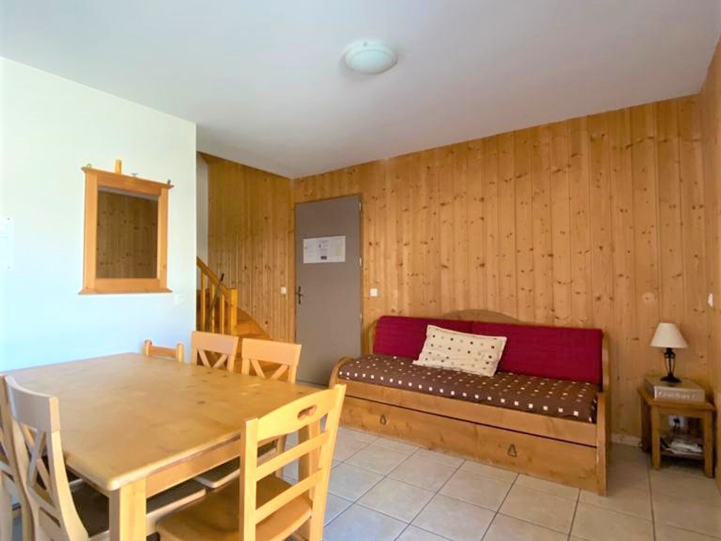 A compact room with a wooden floor, a dining table with chairs, a red sofa, and a small bedside table with a lamp.