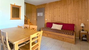 Una habitación compacta con suelo de madera, una mesa de comedor con sillas, un sofá rojo y una pequeña mesita de noche con lámpara.