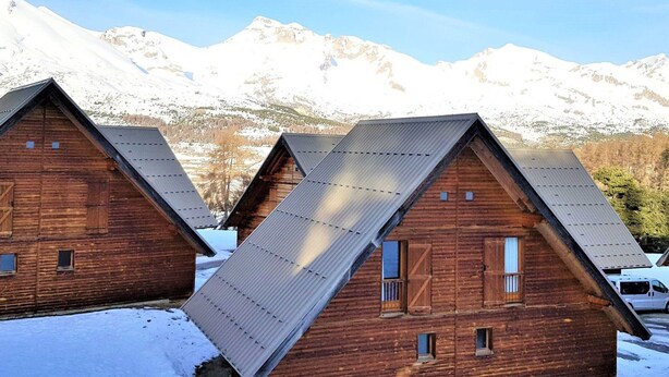 A row of wooden cabins with metal roofs, set against a backdrop of snow-covered mountains.