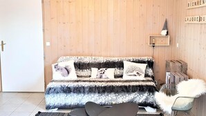 A cozy room with a wooden wall, a sofa with patterned cushions, a fluffy chair, and a small shelf with a decorative item.