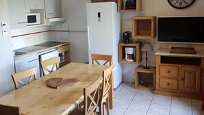 Une cuisine compacte avec des meubles blancs, un four à micro-ondes, une cuisinière et un réfrigérateur. On y trouve une table à manger en bois avec des chaises, une horloge au mur et un téléviseur posé sur un meuble en bois.