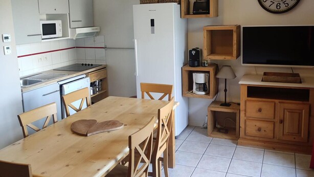 A compact kitchen with white cabinets, a microwave, a stove, and a refrigerator. There is a wooden dining table with chairs, a clock on the wall, and a television on a wooden stand.