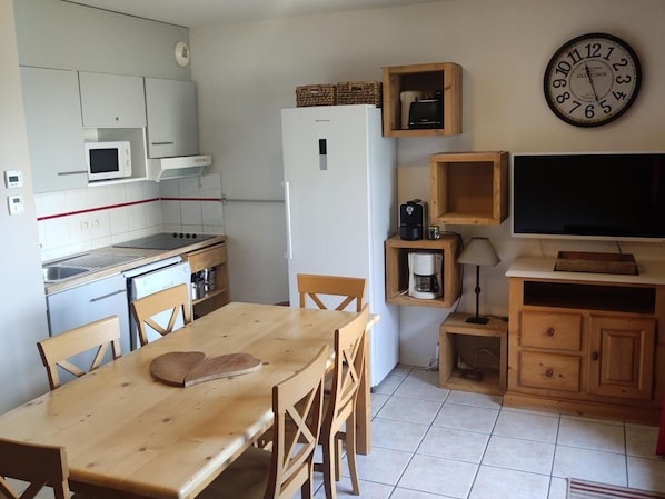 Eine kompakte Küche mit weißen Schränken, einer Mikrowelle, einem Herd und einem Kühlschrank. Es gibt einen Holztisch mit Stühlen, eine Uhr an der Wand und einen Fernseher auf einem Holzschrank.