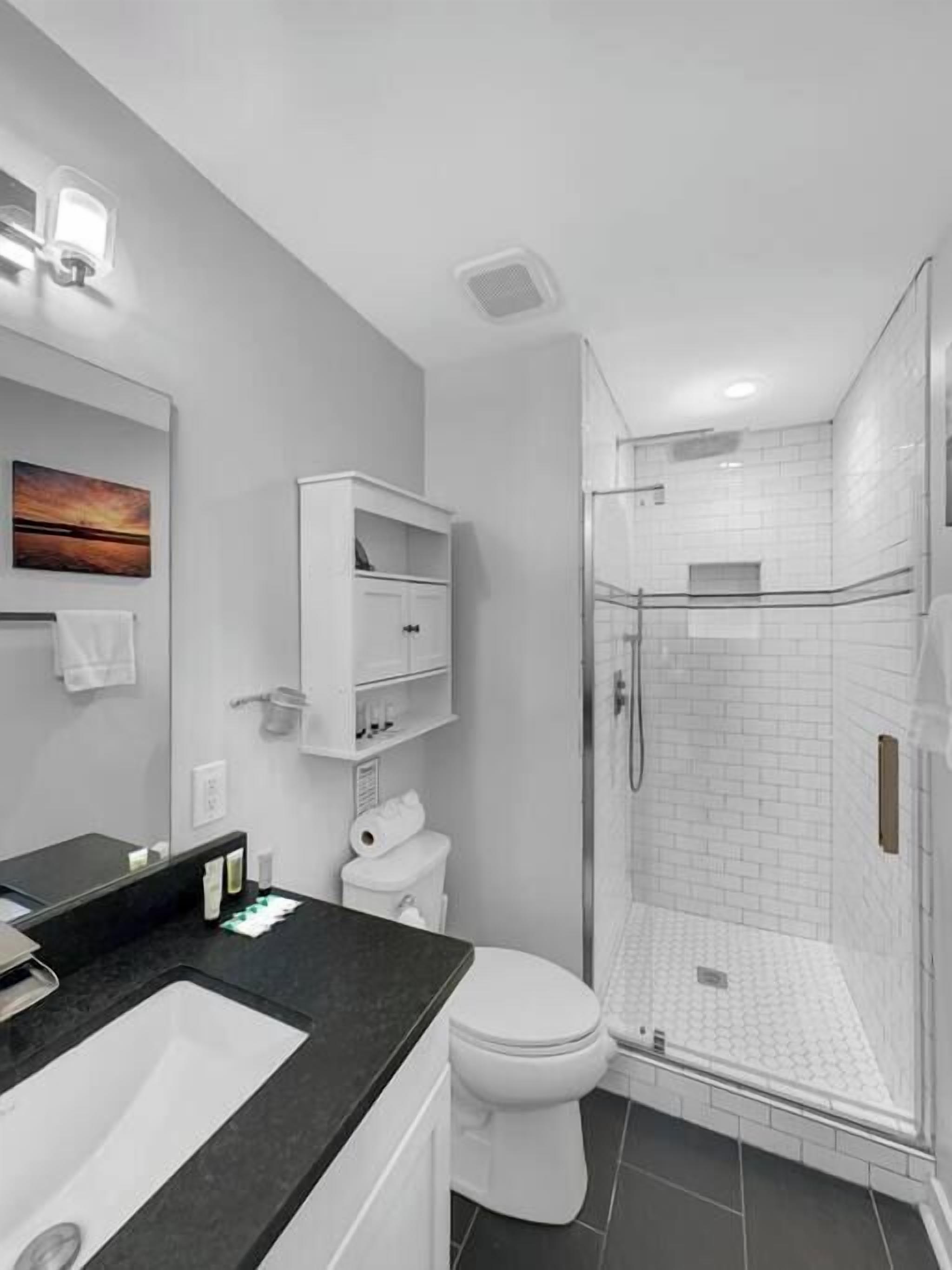 Un baño moderno con lavamanos de superficie negra, inodoro blanco, ducha con mampara de vidrio y armario blanco con estantes.