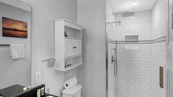 Un baño moderno con lavamanos de superficie negra, inodoro blanco, ducha con mampara de vidrio y armario blanco con estantes.