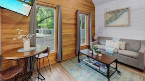 A cozy living room with a wooden ceiling, a sofa, a coffee table, and a TV displaying a beach scene. - Private Hot Tub Island Cottage Near Folly Beach (Charleston)
