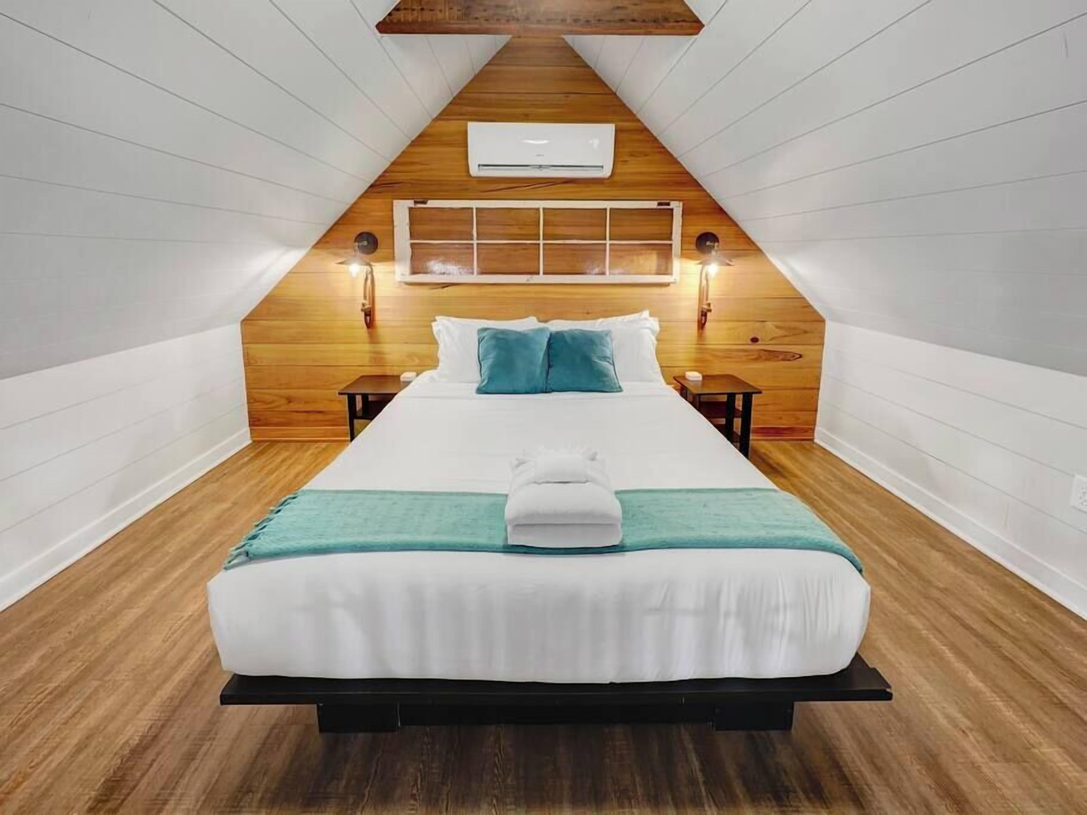 Une chambre avec un plafond en pente en bois, un lit avec une literie blanche et des touches de couleur turquoise, deux tables de chevet et un climatiseur fixé au mur.