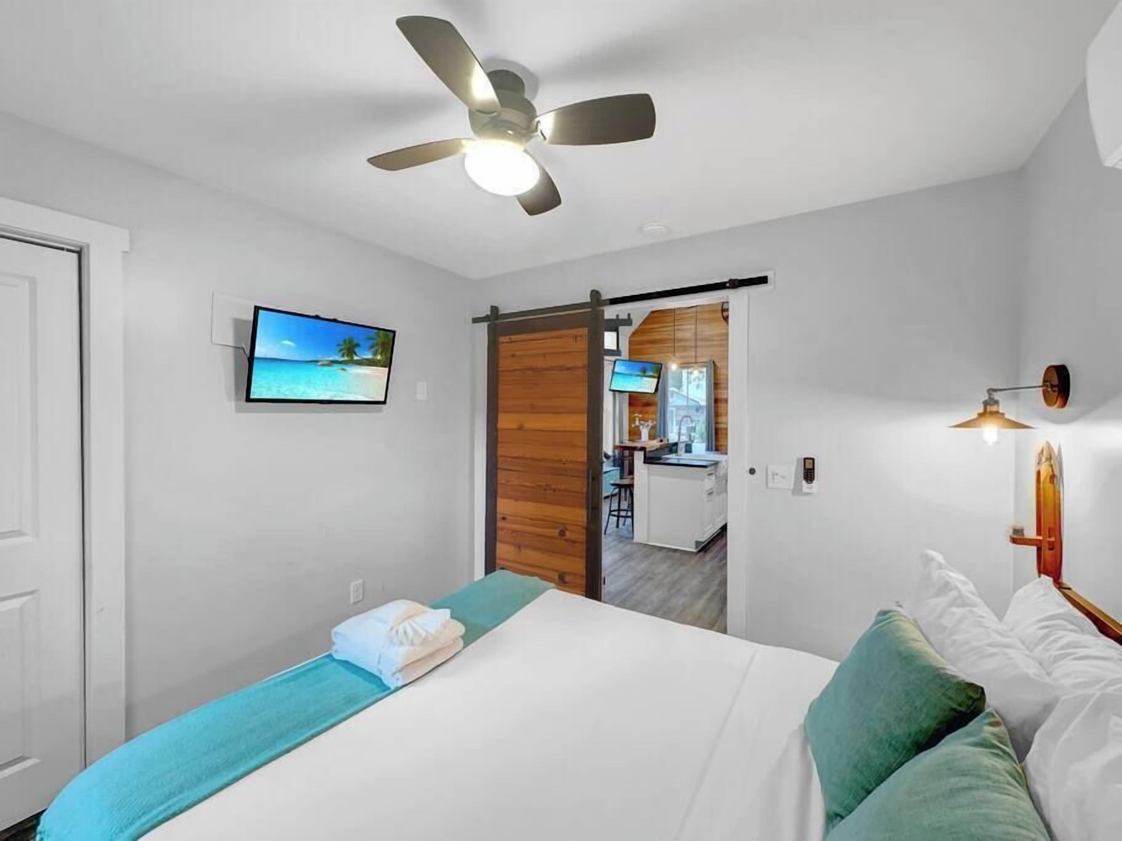 Ein Schlafzimmer mit Bett, Deckenventilator, Fernseher und einer Schiebetür zu einem Küchenbereich.