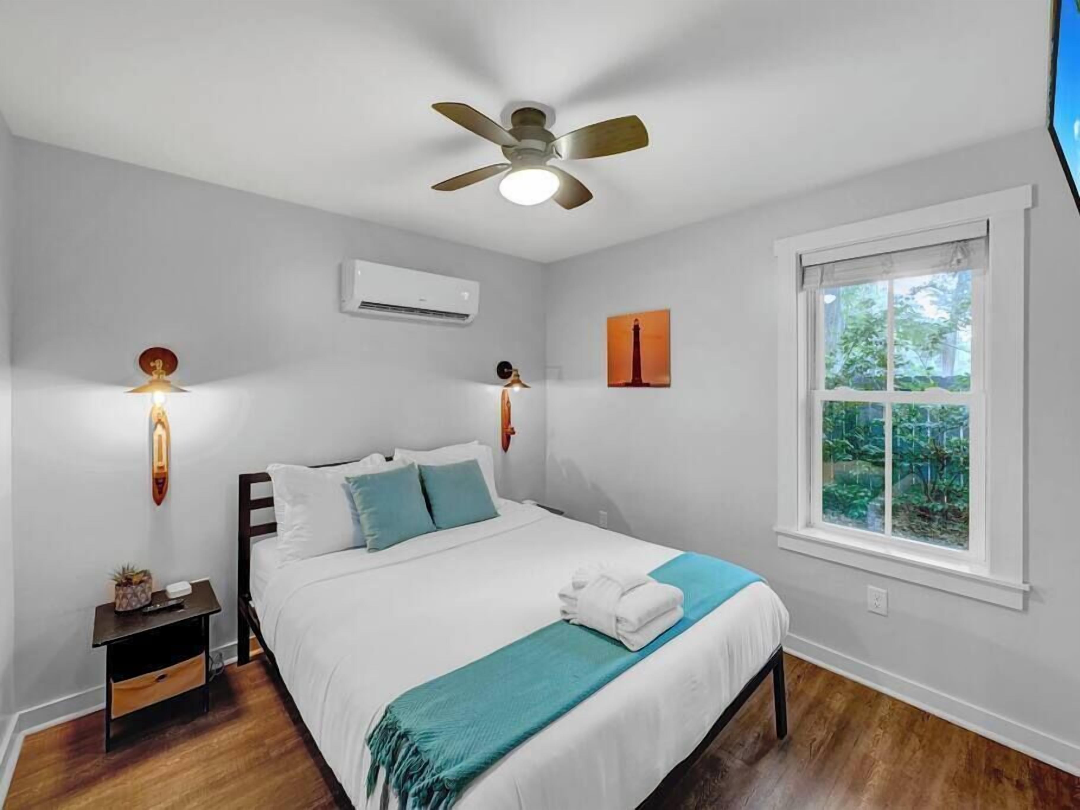 Un dormitorio con cama, ventilador de techo, unidad de aire acondicionado, mesita de noche, ventana con vistas a zonas verdes e iluminación en la pared.