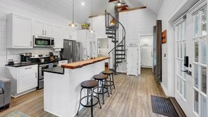 A modern kitchen with white cabinets, a central island with stools, a spiral staircase, and a large window. - Private Hot Tub Island Cottage Near Folly Beach (Charleston)