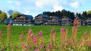 A row of wooden chalet-style buildings with balconies, set against a backdrop of mountains and a clear sky. - Family chalet in La Joue du Loup with parking (La Joue du Loup)