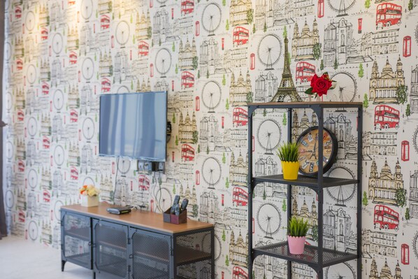 A room with a patterned wallpaper, a TV mounted on the wall, a wooden cabinet, and a black shelving unit with plants. - A Luxurious & Comfortable Home Near USM & Spice (Desa Mutiara Ii)