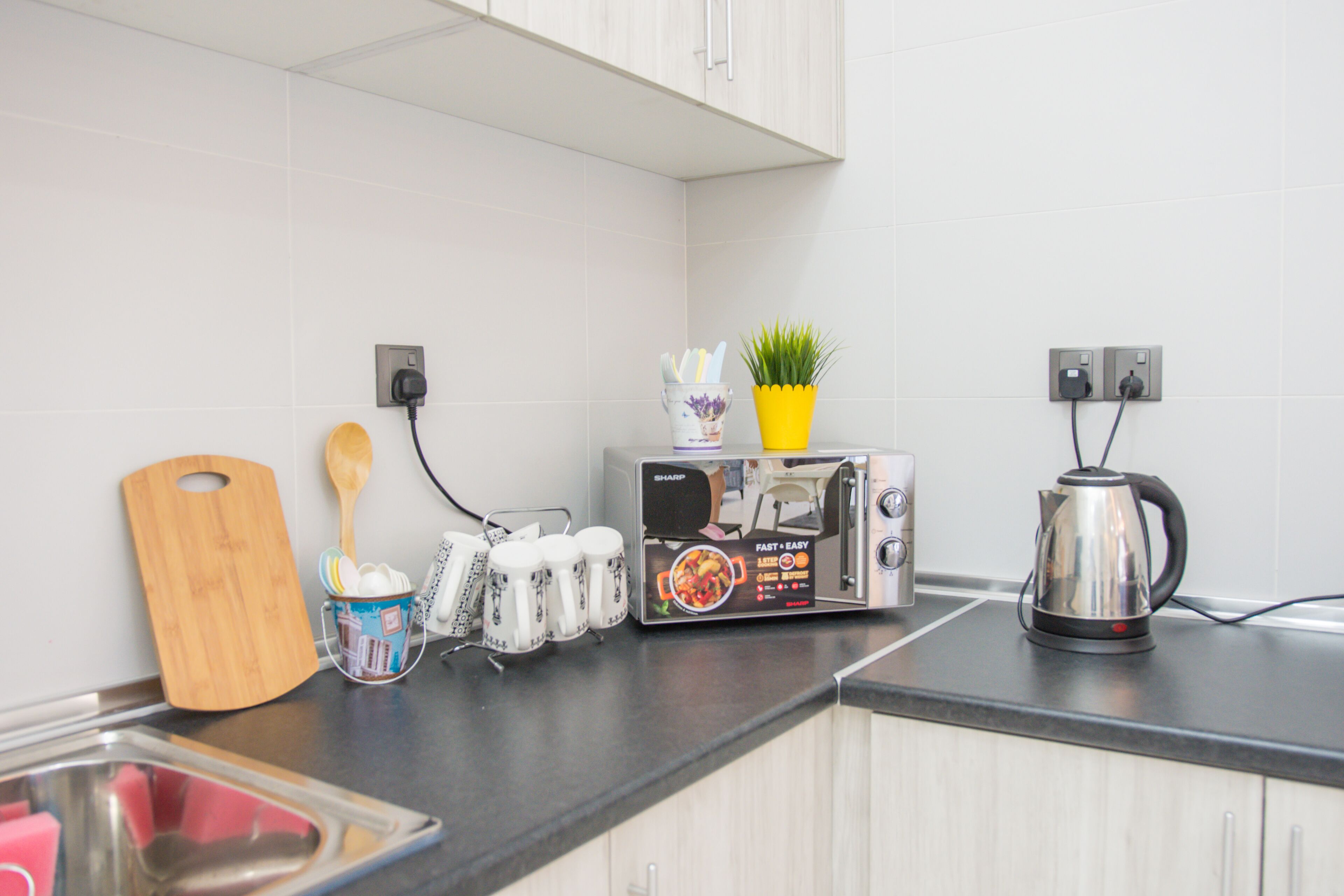 A kitchen with a toaster, a kettle, a wooden cutting board, a plant, and a cup holder with utensils.