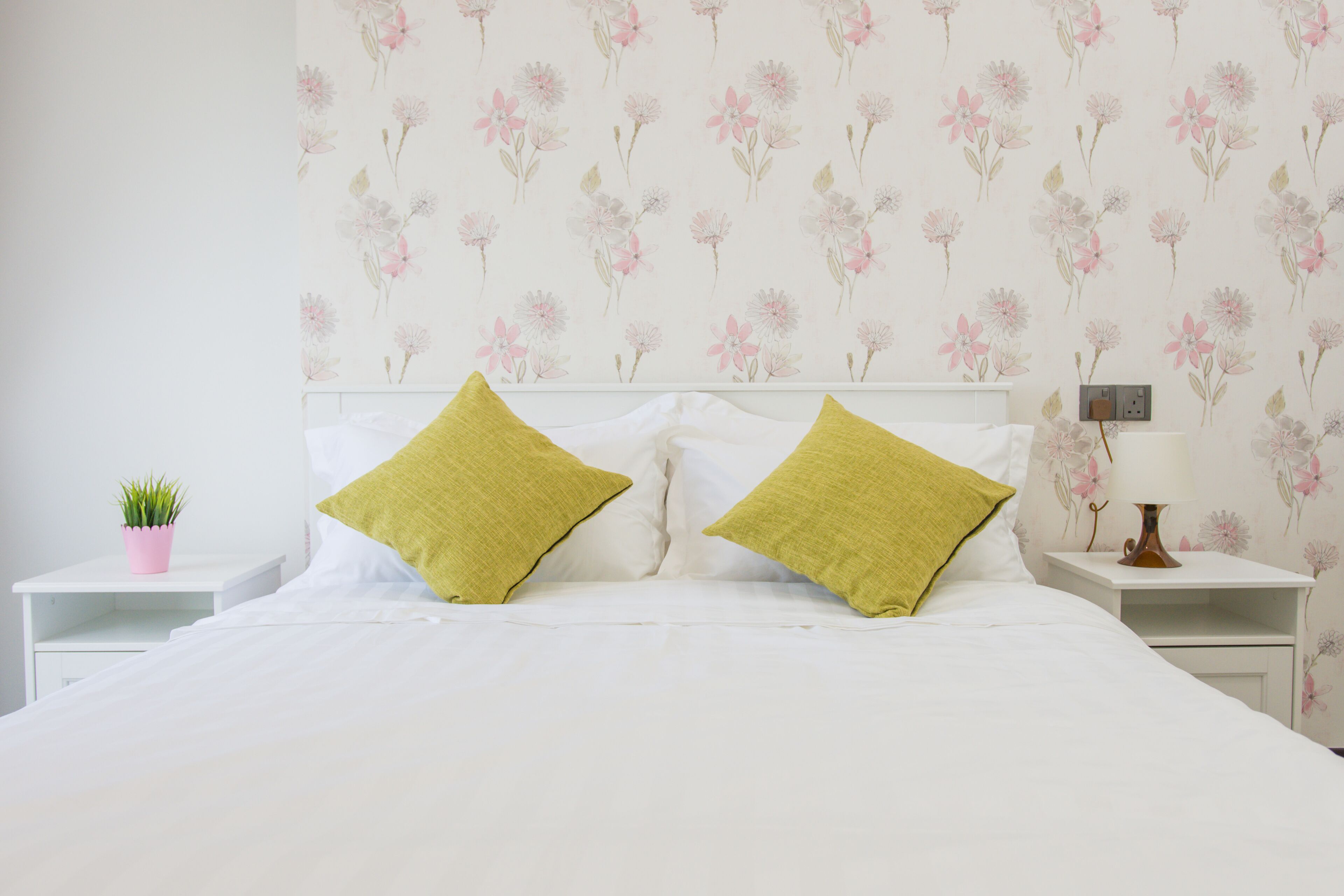 A neatly made bed with white linens and two green pillows, a small pink plant on a white nightstand, and a wall with a floral pattern.
