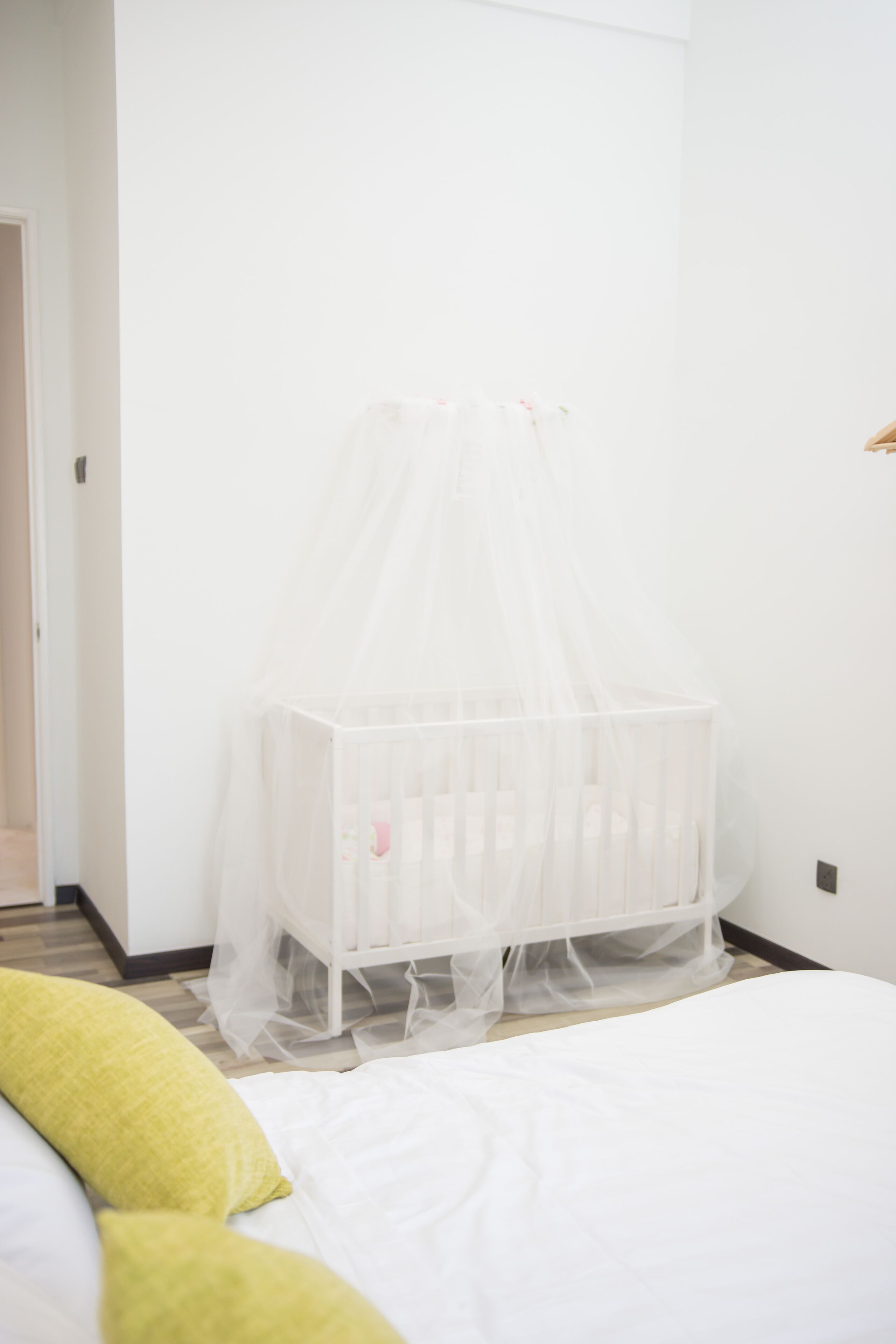 Un berceau blanc surmonté d’un dais transparent, dans une pièce aux murs blancs et où se trouve un lit recouvert de linge de lit blanc.
