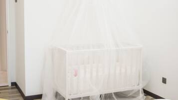 Un berceau blanc surmonté d’un dais transparent, dans une pièce aux murs blancs et où se trouve un lit recouvert de linge de lit blanc.