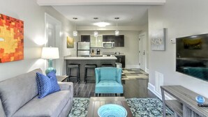 A modern living room with a grey sofa, a blue armchair, and a coffee table. A kitchen is visible in the background with a microwave, stove, and bar stools. - Charming Historic King James Building Unit #4 (Asheville)