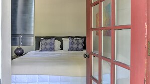 A neatly made bed with patterned pillows, a bedside lamp, and a window with blinds. - Charming Historic King James Building Unit #4 (Asheville)