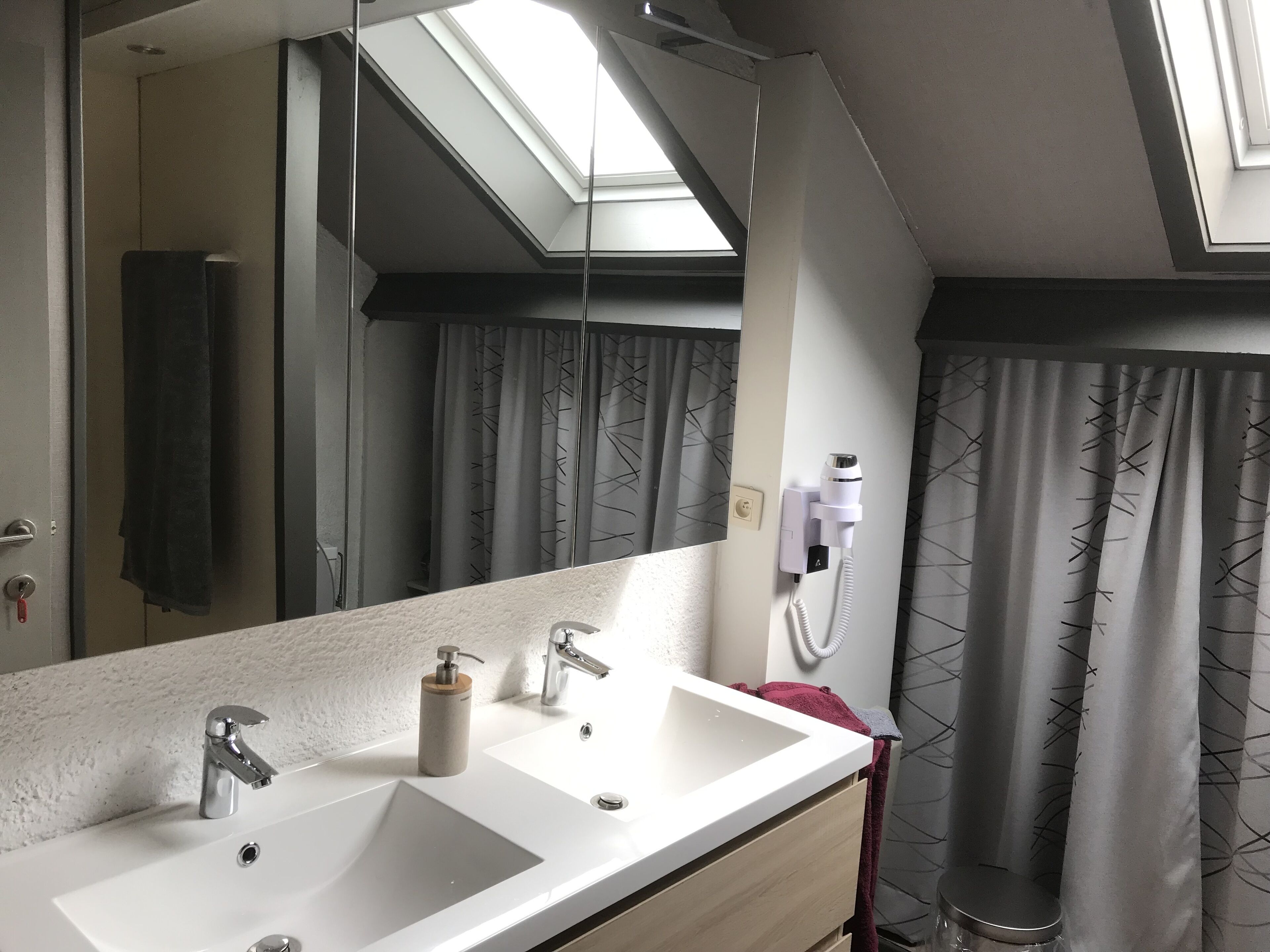 Une salle de bain avec un grand miroir, deux lavabos, un porte-serviettes, un rideau de douche et un luminaire au plafond.