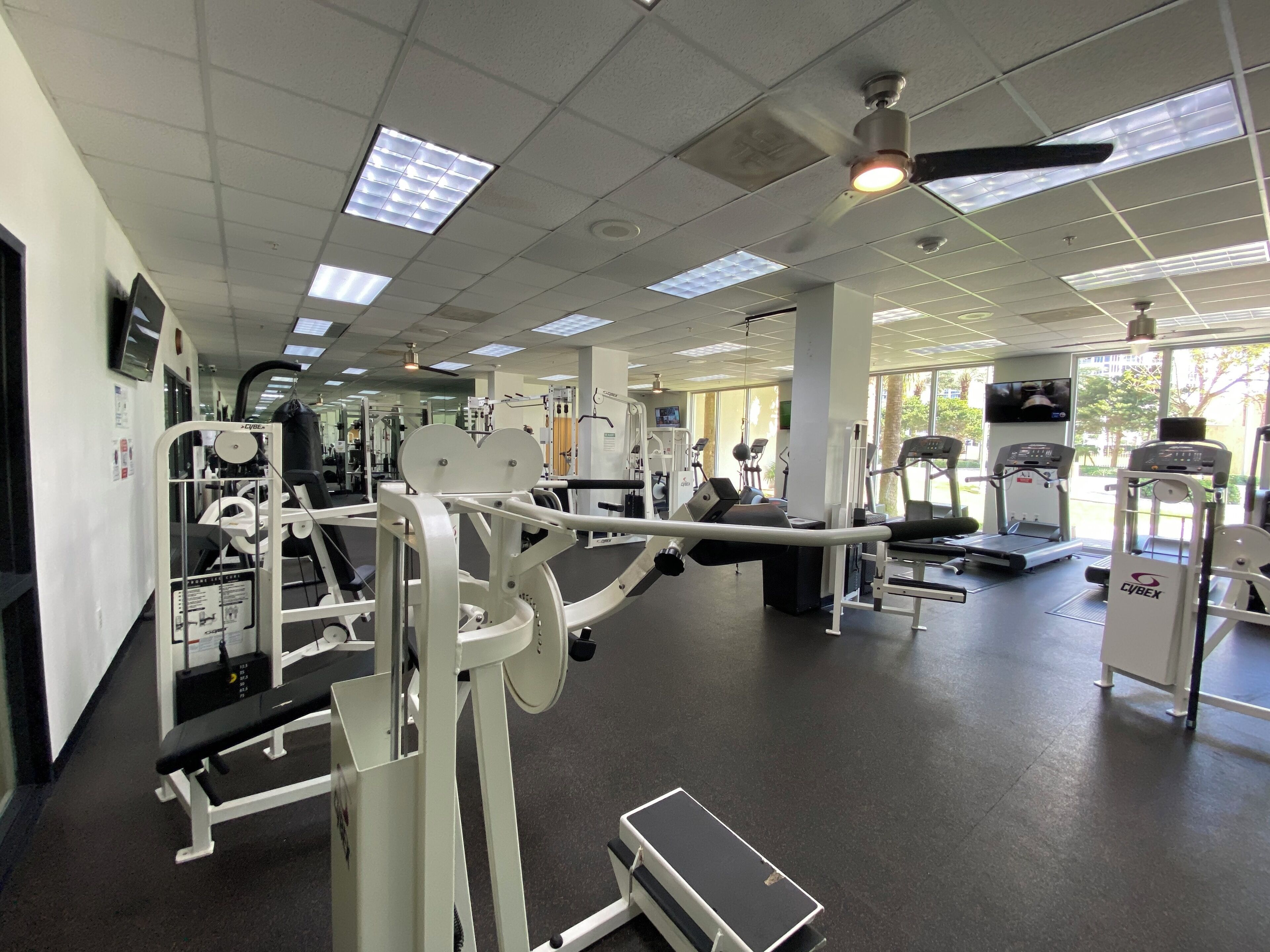 Een sportschool met diverse fitnessapparaten, een loopband en een plafondventilator.