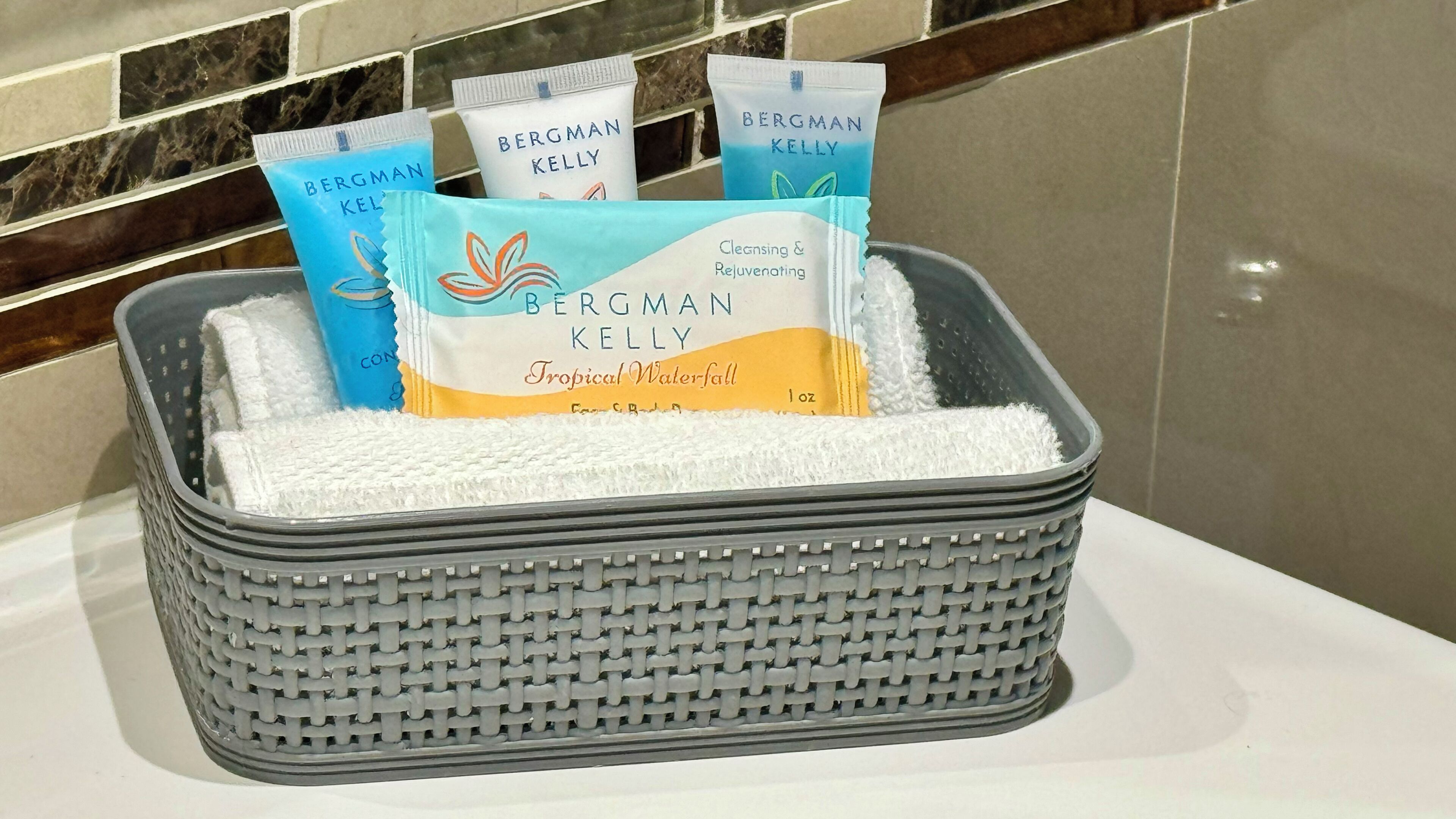 Un panier tressé gris contenant plusieurs produits nettoyants et régénérants de la marque Bergman Kelly, dont le produit Tropical Waterfall, posé sur le comptoir d’une salle de bain.