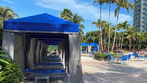 A covered outdoor seating area with blue chairs and tables, surrounded by palm trees and a tall building in the background. - SUNNY ISLES CONDO-RESORT (PARKING INCLUDED) (Sunny Isles Beach)