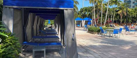 A covered outdoor seating area with blue chairs and tables, surrounded by palm trees and a tall building in the background.