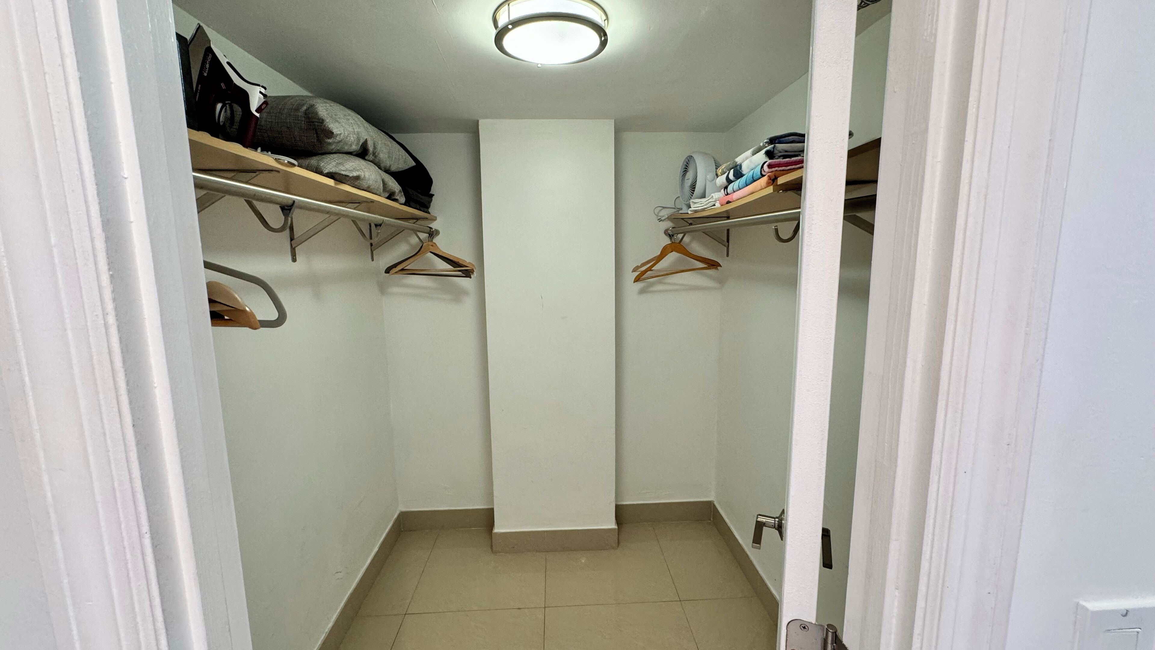 A narrow walk-in closet with wooden shelves and hangers, a white door, and a tiled floor.