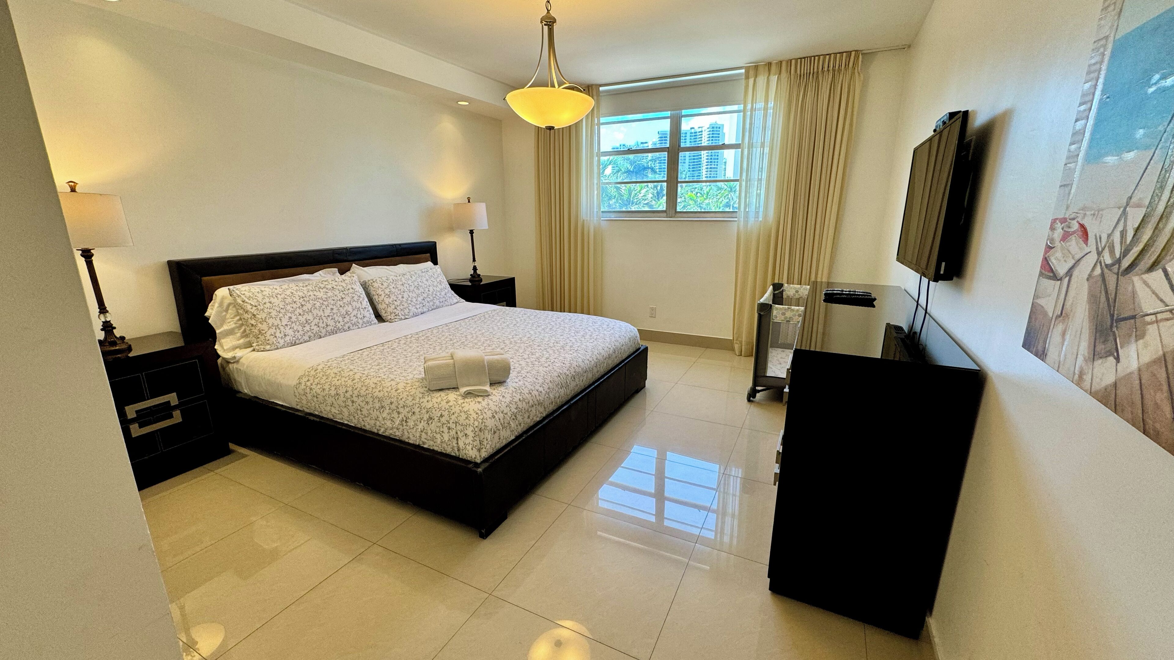A hotel room with a large bed, bedside lamps, a window with curtains, a flat-screen TV mounted on the wall, and a small desk with a chair.